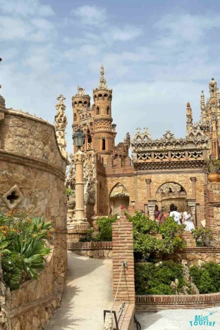 A detailed, whimsical brick and stone castle with ornate towers and lush greenery, set against a partly cloudy sky. People walk along the path in front.