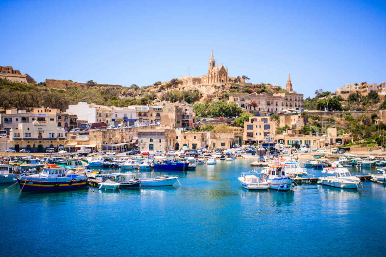 Colorful boats docked in a clear blue harbor with buildings and a hillside church in the background under a bright sky.