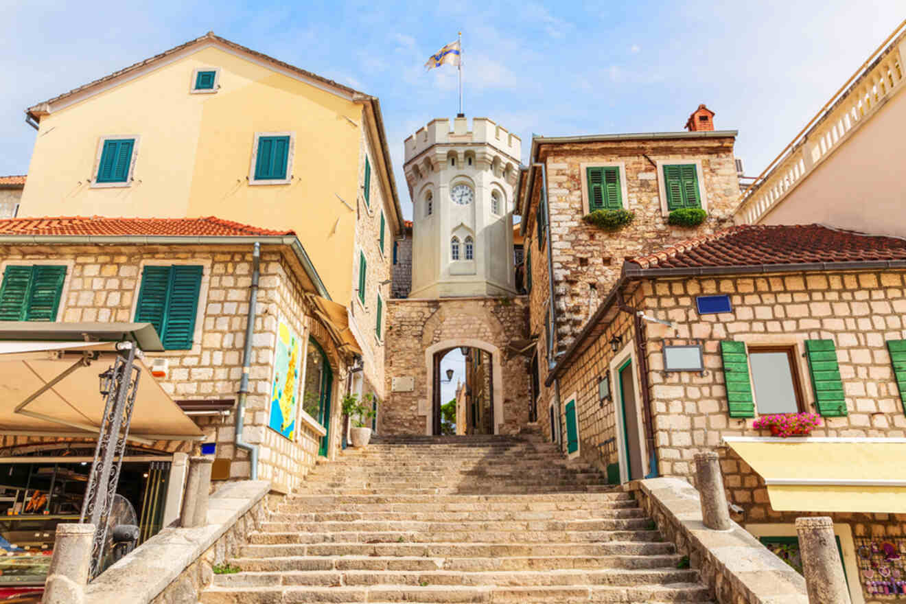 Stone staircase leading up to an arched tower between two rustic buildings with green shutters and a flag on top.