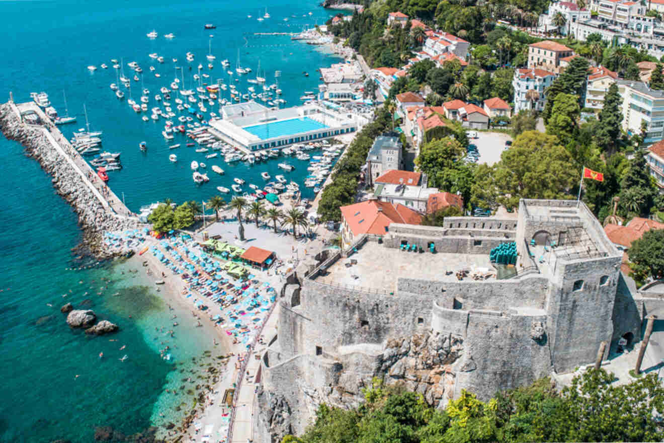 Aerial view of a coastal town with a historic fortress, beach, and marina filled with boats. Surrounding area includes residential buildings and lush greenery.