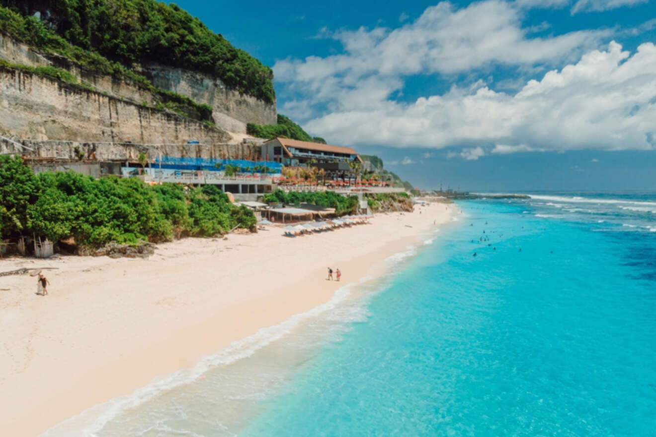 Beach with white sand and clear blue water, lined by cliffs and greenery. A beachside building and umbrellas are visible. People are swimming and walking along the shore under a partly cloudy sky.