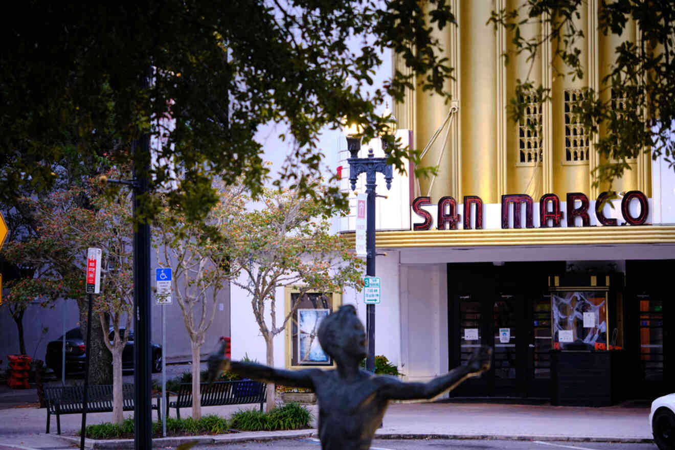 Art deco theater with "San Marco" sign, surrounded by trees and a statue in the foreground.