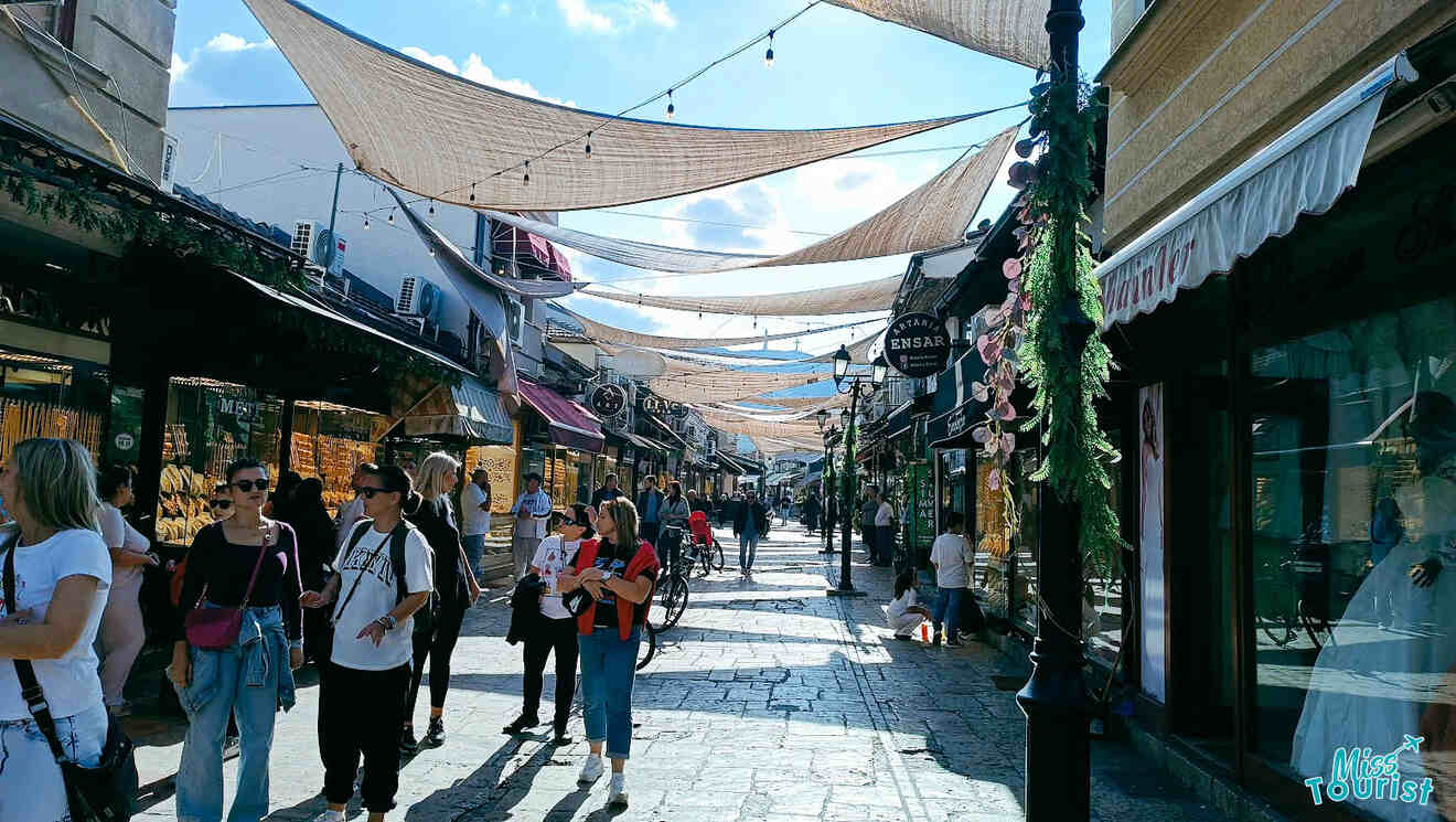 A busy street market with people walking and shopping. Canopies provide shade overhead, and various shops line both sides of the cobblestone path.