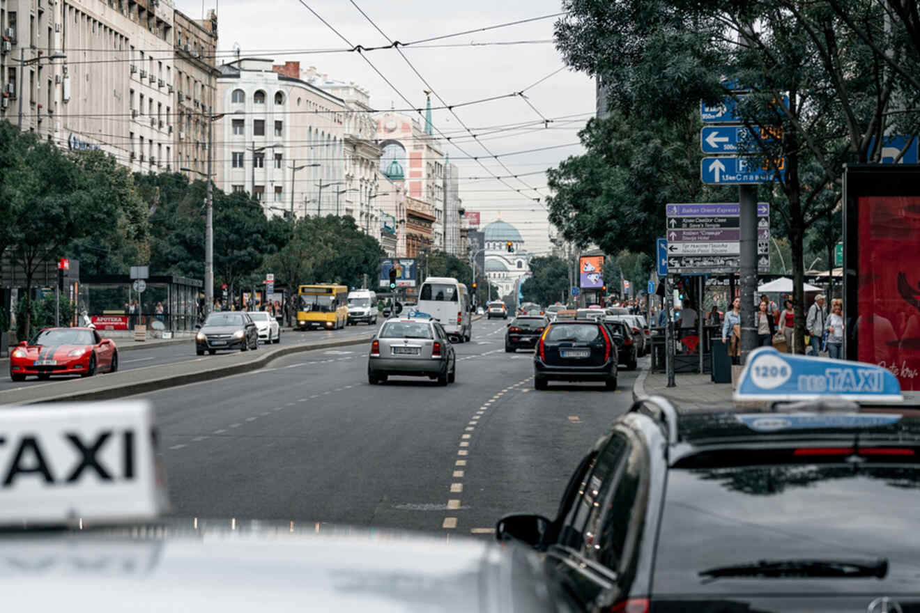 City street scene with multiple vehicles and pedestrians. Buildings line the road, and signage is visible. The sky is overcast.