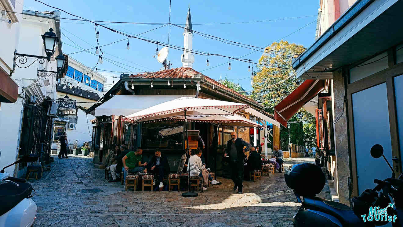 Outdoor café with people sitting under umbrellas in a cobblestone plaza. A tall minaret is visible in the background, surrounded by buildings and string lights.