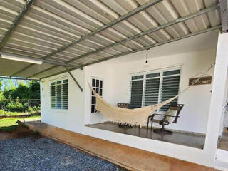 A white house with a covered porch features a hammock and two chairs. The area is paved with gravel and surrounded by greenery.