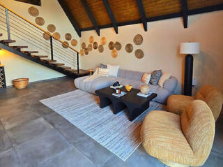 Modern living room with a gray sectional sofa, two mustard armchairs, a black coffee table, and a large white rug. Wicker baskets decorate the wall and stairs feature a metal railing.