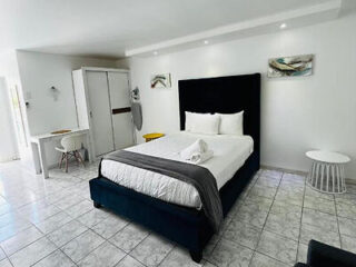 A neatly made bed in a modern, minimalist white bedroom with a desk, chair, wardrobe, and decorative wall art.