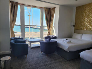 Hotel room with a bed, three blue chairs, a table, and a sea view through large windows with beige curtains.