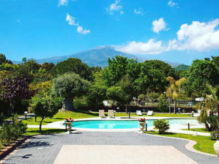 A serene garden features a circular blue pool, surrounded by lush greenery and trees under a bright blue sky with clouds. A mountain is visible in the distance.