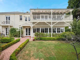 Two-story house with a covered upper balcony, white exterior, and a red brick pathway leading through a green garden.