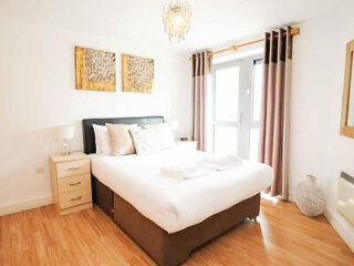 Bright bedroom with a double bed, white bedding, two decorative wall pieces, wooden floor, bedside drawer, and a window with curtains. A chandelier hangs from the ceiling.