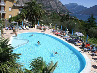 Outdoor pool with people swimming and lounging on chairs. Surrounded by palm trees, buildings, and mountains, with a lake visible in the background under a clear blue sky.