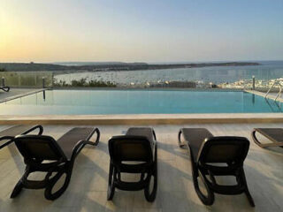 Three empty loungers face an infinity pool overlooking a harbor with boats. The sky is clear at sunset.