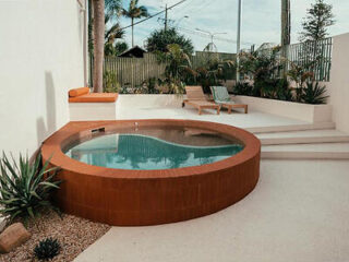 A round outdoor pool with terracotta tiles is surrounded by white walls, greenery, and two lounge chairs on a raised platform.