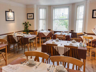 A dining room with several tables and wooden chairs, set for a meal with plates, cups, and cutlery. Natural light streams through large windows with vertical blinds.