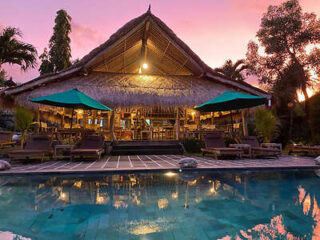 A tropical, thatched-roof pavilion with green umbrellas sits beside a clear swimming pool, set against a pink sunset sky.