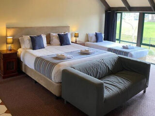 Hotel room with two beds, a gray sofa, nightstands, lamps, and large window with green landscape view outside. Towels are placed on each bed.