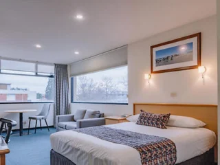 A hotel room with a double bed, two chairs, a small table, and a large window. The wall features a framed picture above the bed.