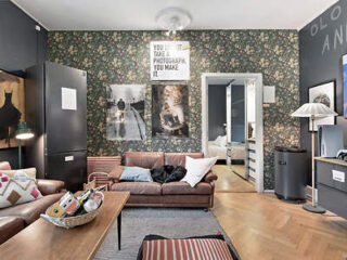 Living room with floral wallpaper, brown leather sofa, patterned cushions, wall art, and wooden coffee table. Doorway leads to a bedroom with visible bed. Floor lamp and storage units present.