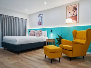 Modern bedroom with a large bed, teal accent wall, curtains, and a yellow armchair with a footrest. A standing lamp and potted plant are nearby. Two framed pictures hang on the wall.