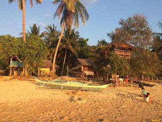 A beach scene with a wooden outrigger boat on the sand, surrounded by palm trees and rustic huts under a clear blue sky.