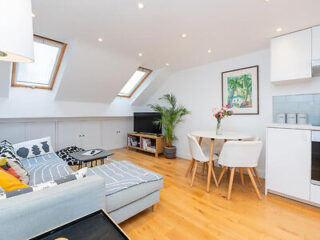 A bright, modern living room with skylights, a corner sofa, a TV, a small dining table with chairs, and a potted plant on wooden flooring.