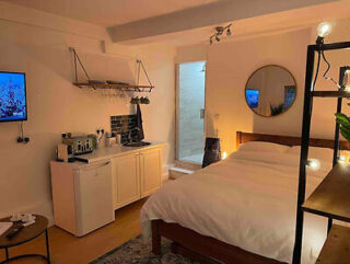 Cozy bedroom with a bed, round mirror, kitchenette, and open bathroom door. Warm lighting enhances the ambiance.