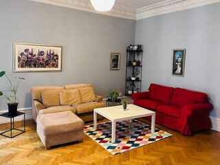 A living room with a beige sofa, red couch, white coffee table on a colorful rug, and two paintings on gray walls. A potted plant is in the corner.