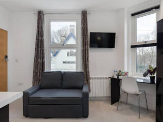 A small room with a gray sofa, wall-mounted TV, and a desk with a chair. Two windows have dark curtains. A white radiator and some plants are visible.