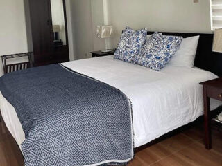 A neatly made bed with white sheets, two decorative pillows, and a patterned dark blue throw. Flanked by wooden nightstands, with a wardrobe and lamp visible in the background.