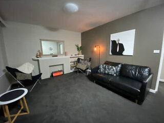 Modern living room with a black leather sofa, butterfly chair, small round table, wall-mounted artwork, and a decorative fireplace. A tall lamp and shelf complete the space.