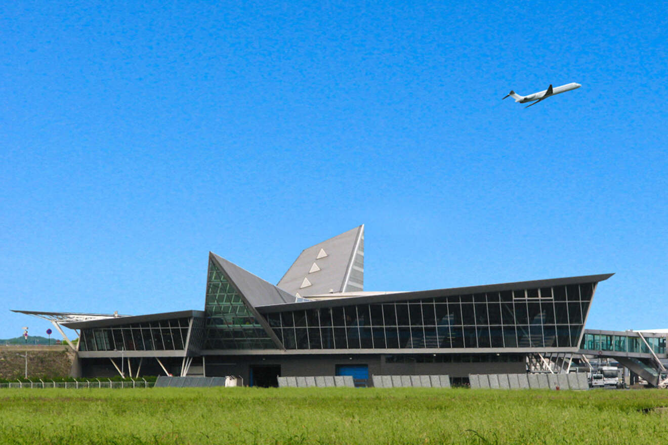 Modern angular airport terminal with a large glass facade, under a clear blue sky. An airplane is taking off in the background.