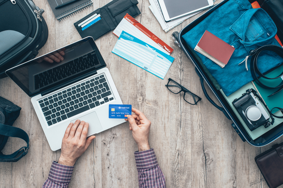 A person using a laptop at a wooden table, surrounded by travel items, including an open suitcase, a camera, glasses, tickets, and a credit card.