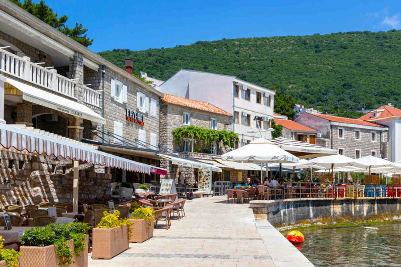 A picturesque street with stone buildings, outdoor cafe seating under umbrellas, and a view of a lush green hillside.