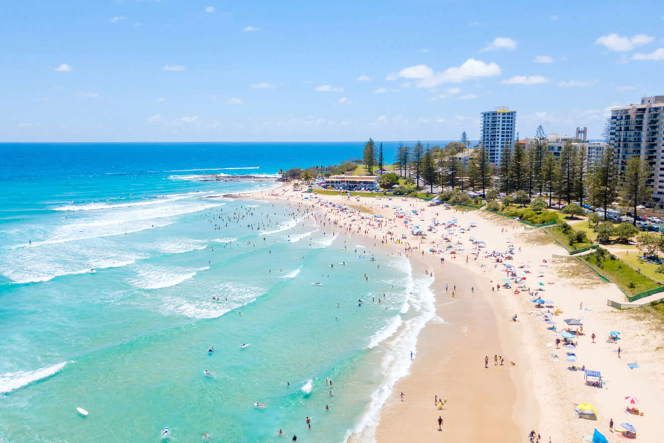 Aerial view of a crowded beach with people swimming and sunbathing. Blue ocean waves and high-rise buildings are visible in the background.