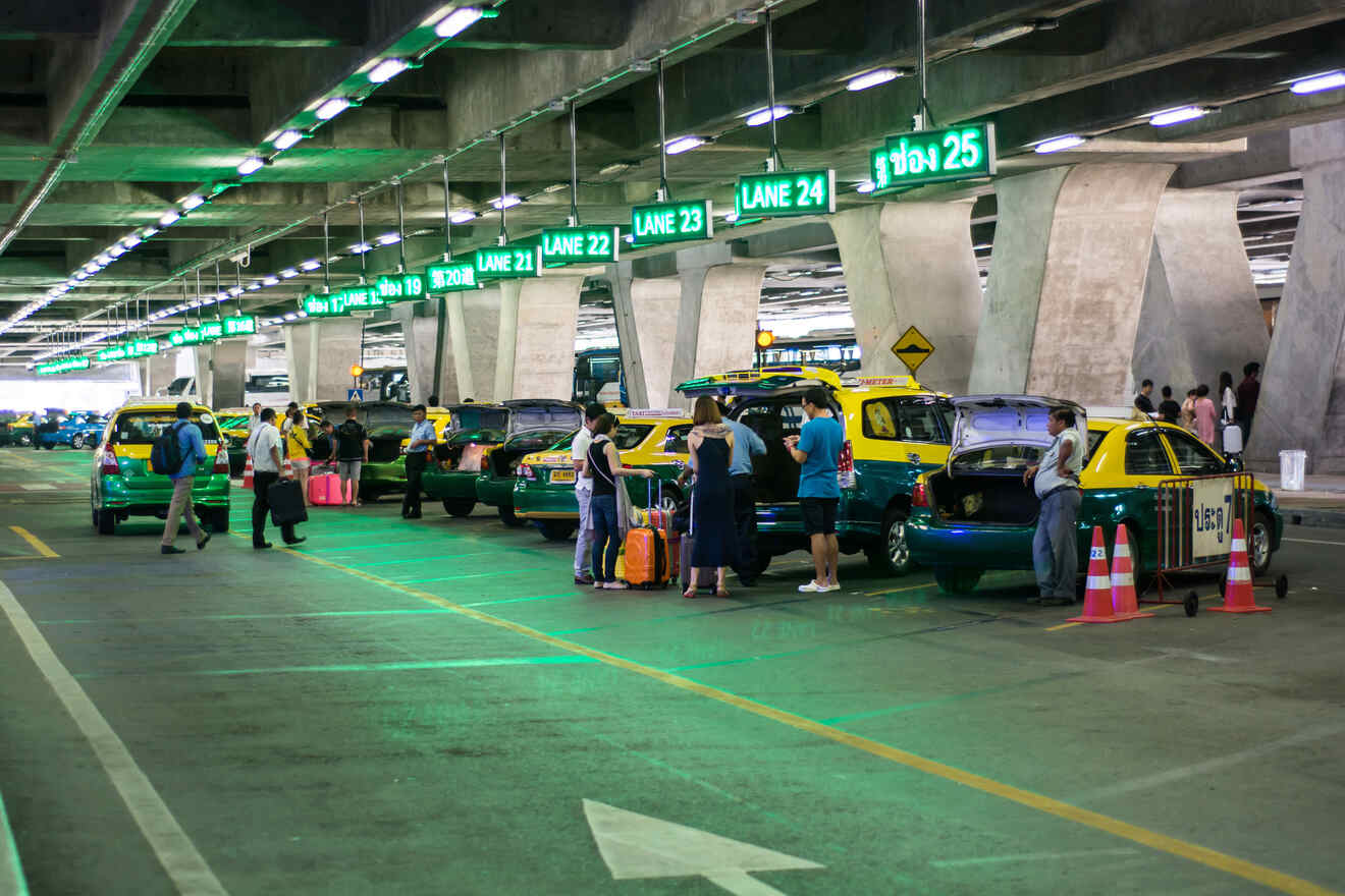 People loading luggage into taxis in an airport parking area beneath concrete beams, with green lane signage illuminated above.