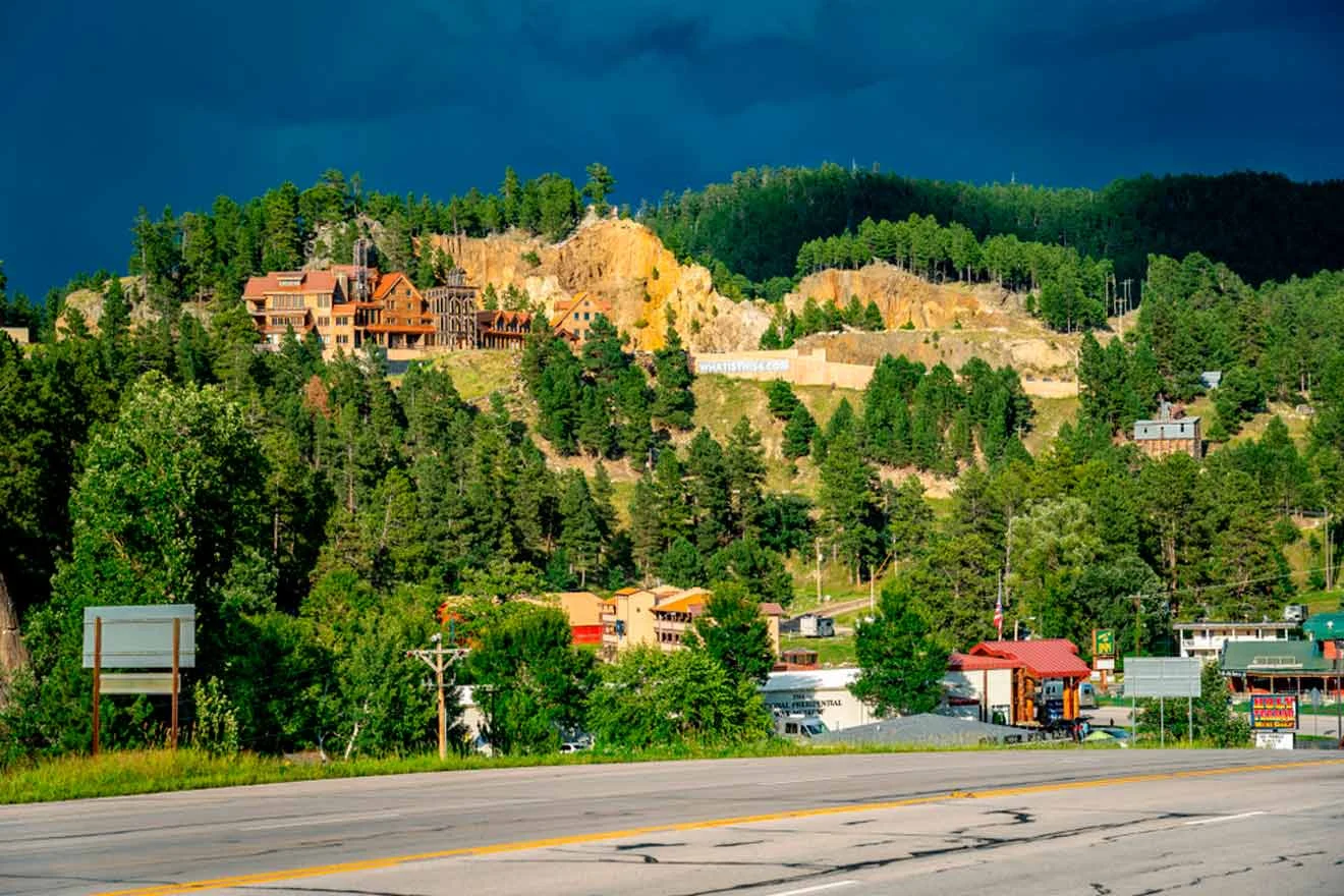 A hilltop with buildings is surrounded by dense trees under a dark, cloudy sky. A roadway and more buildings are visible in the foreground.