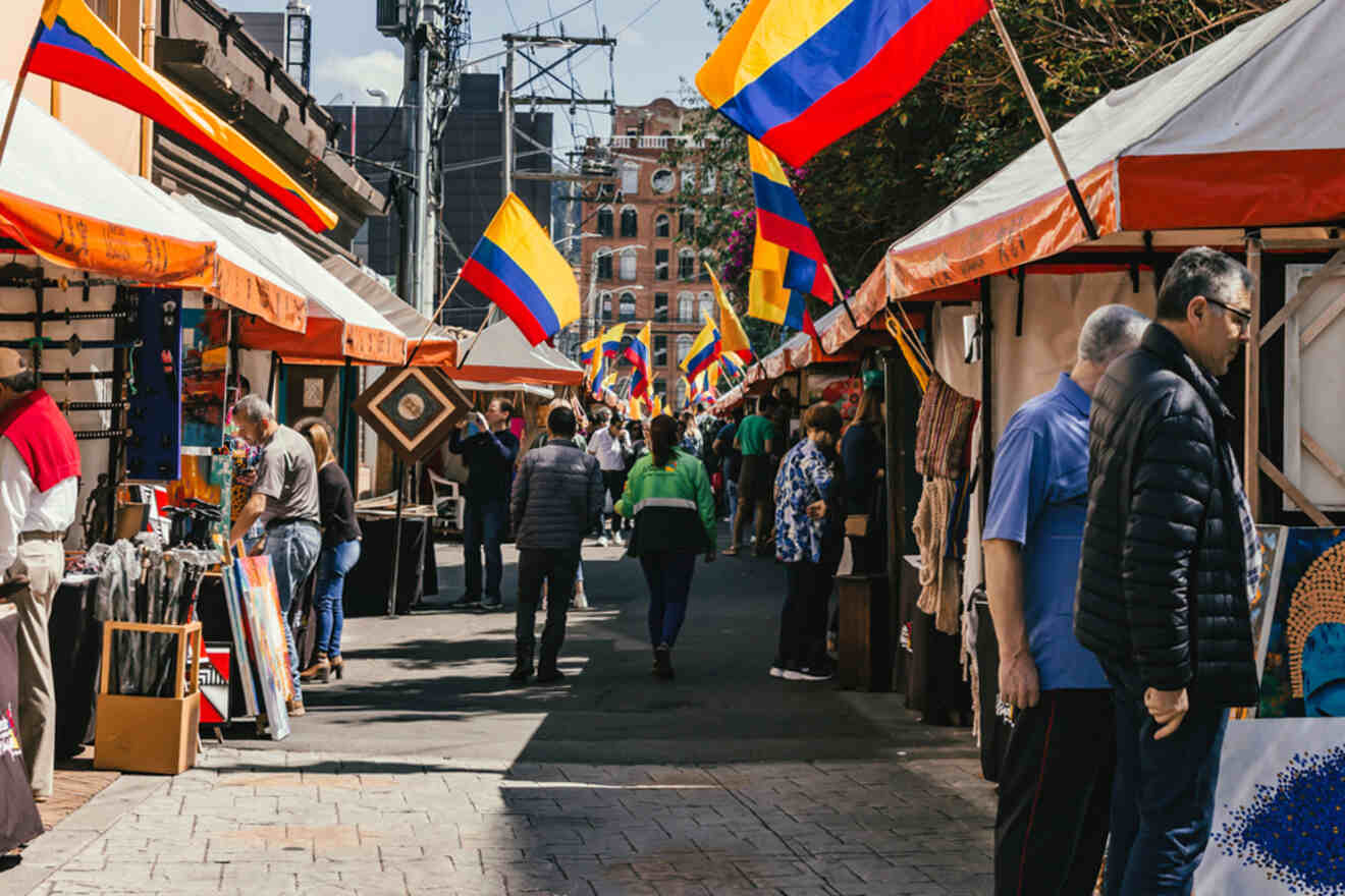 Outdoor market with people walking and browsing stalls. Colombian flags are displayed above the tents. The setting is urban, with buildings and trees in the background.