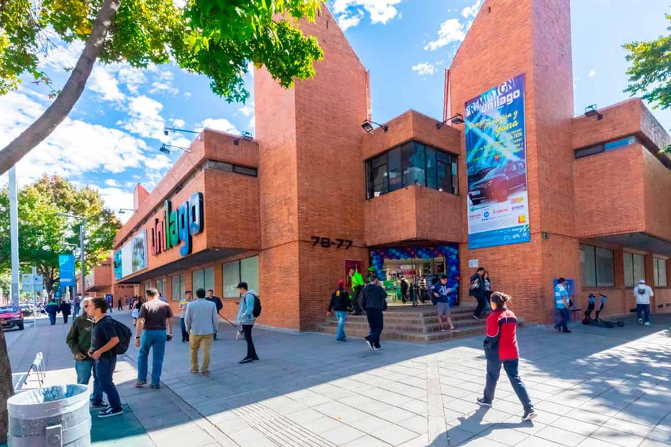 A red brick building with a mall entrance. People are gathered outside. Banners and signs are displayed on the walls. Trees and a blue sky are visible in the background.