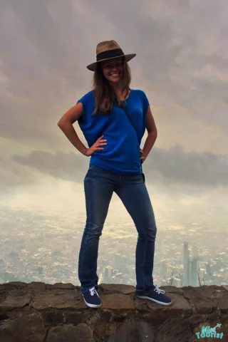 the author of the post with a hat stands on a stone wall, city skyline visible in the background under a cloudy sky.