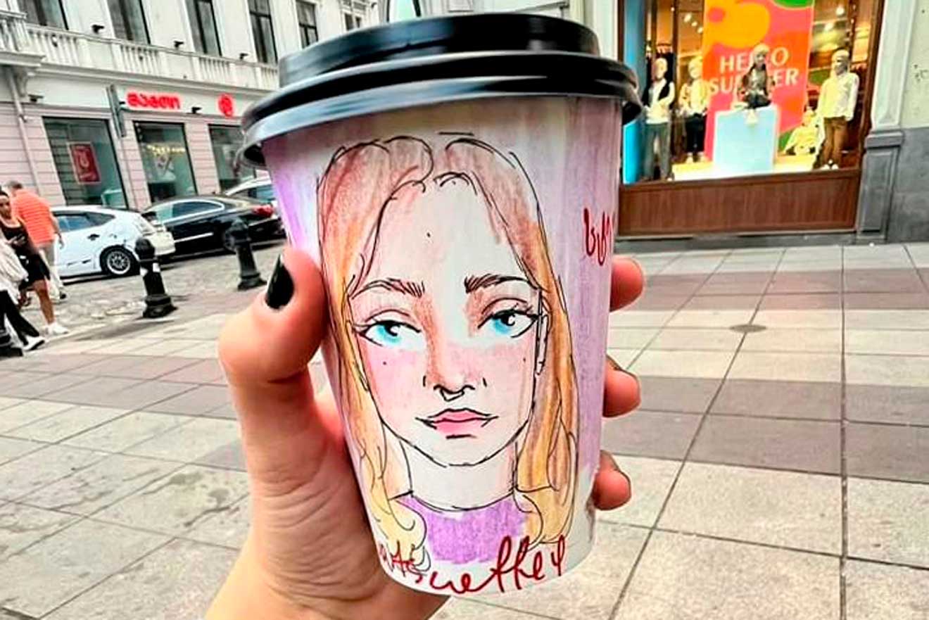 A hand holds a coffee cup with a hand-drawn portrait of a woman. Buildings and pedestrians are visible in the background.