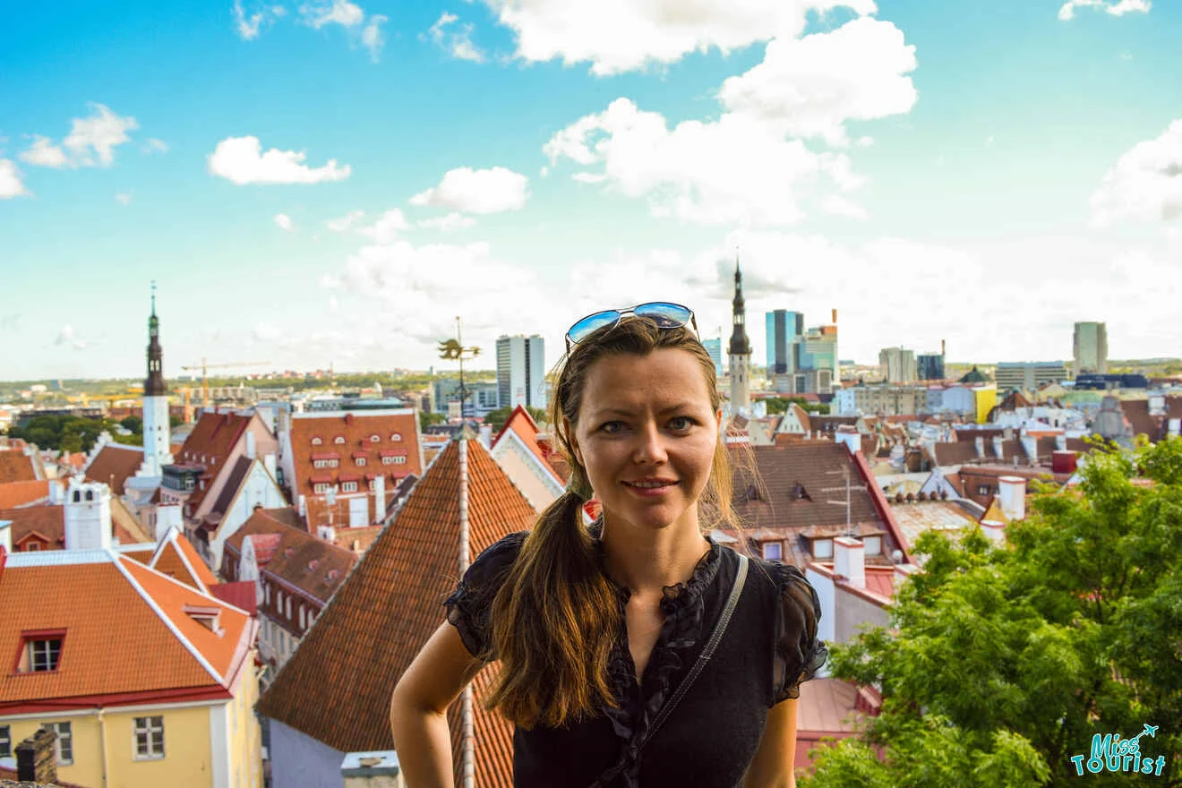 Yulia, the founder of this website, stands smiling in front of a scenic cityscape with red-roofed buildings and a clear blue sky.