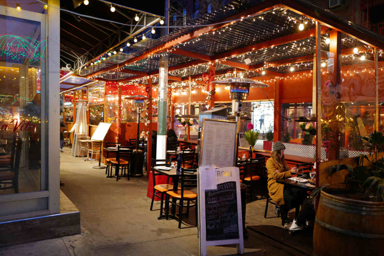 A cozy outdoor restaurant seating area with string lights, red decor, and several tables occupied by patrons. A menu board is visible at the entrance.