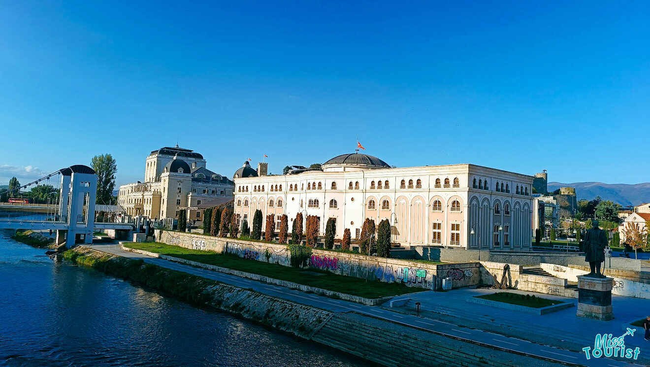 View of a large, neoclassical building by a river under a clear blue sky, with a pedestrian bridge nearby and a statue in the foreground.