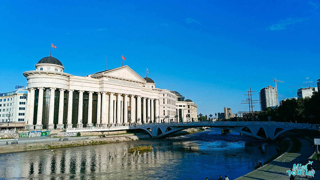 A neoclassical building with columns and domes sits by a river. A white arched bridge spans the water under a clear blue sky.