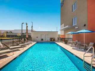 Outdoor swimming pool with blue water, surrounded by chairs, umbrellas, and a building. Clear sky in the background.