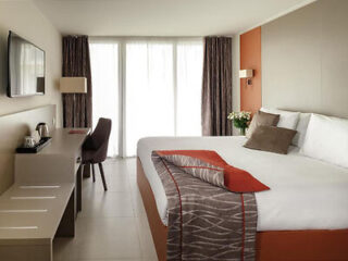 Hotel room with a neatly made double bed, pillows, a desk with a chair, a wall-mounted TV, and a large window with curtains. A red and gray decorative throw is on the bed.