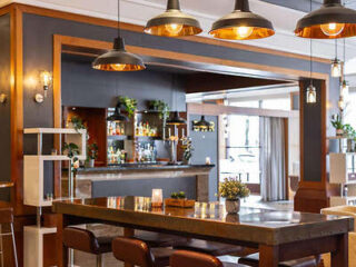 Modern restaurant interior with wooden tables, pendant lights, and a well-stocked bar in the background. Plants and soft lighting add a cozy ambiance.