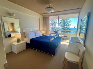 A modern hotel room with a double bed, blue comforter, two side tables with lamps, and a view of the ocean through large glass doors leading to a small balcony with a chair.